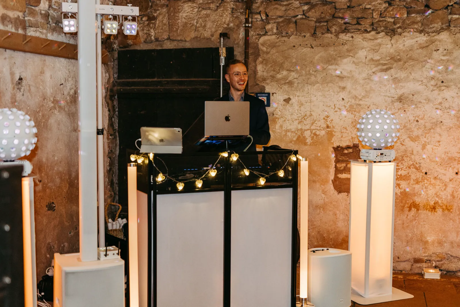 Jannik an seinem weißen DJ-Pult mit Lichterkette vor einer Natursteinwand